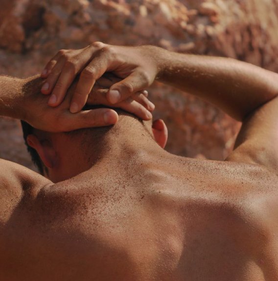 A man's bare back and neck with both hands clasped behind his head, against a rock wall
